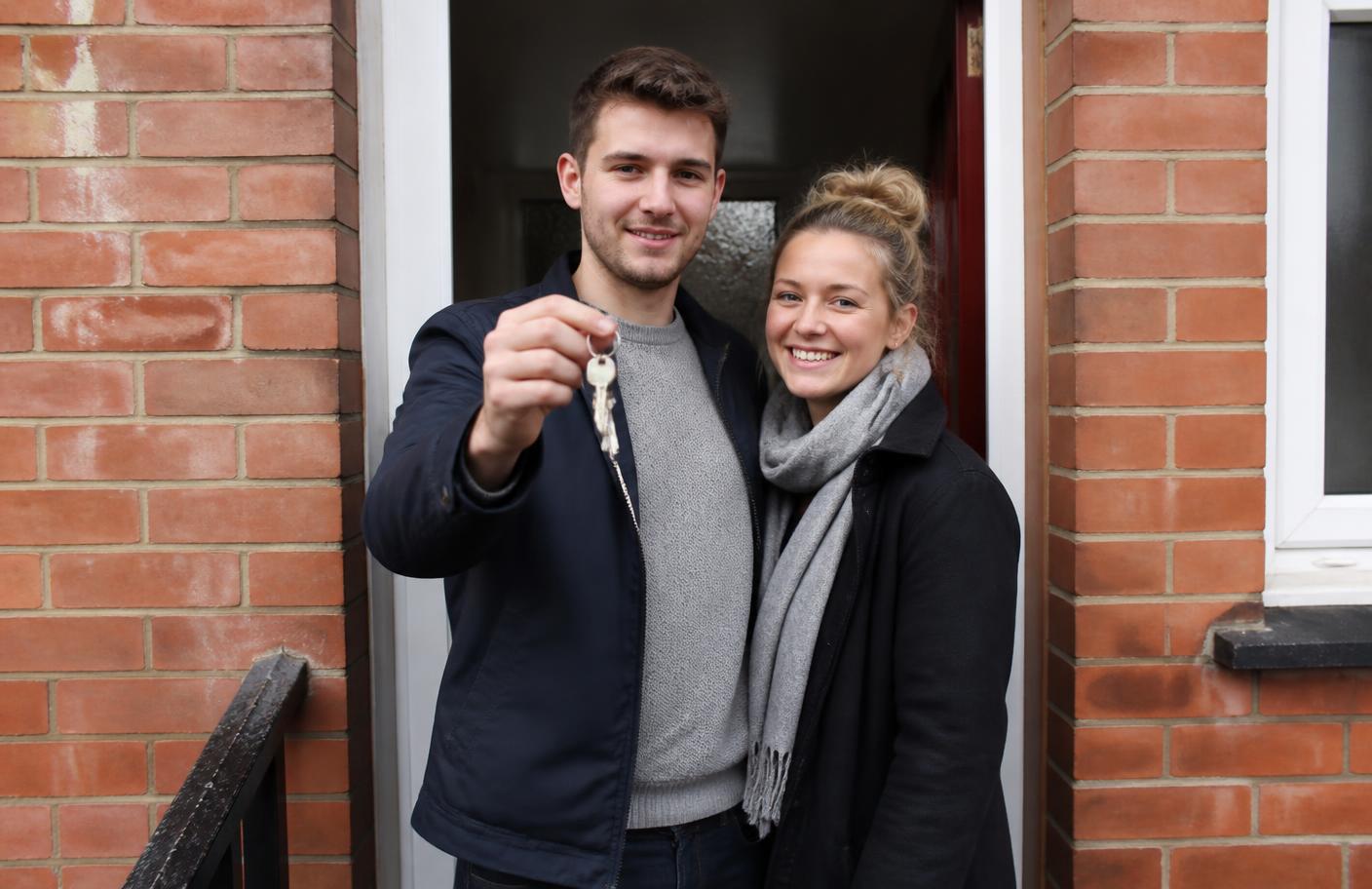 A young couple holding the keys to their first home in the doorway of a UK terraced house